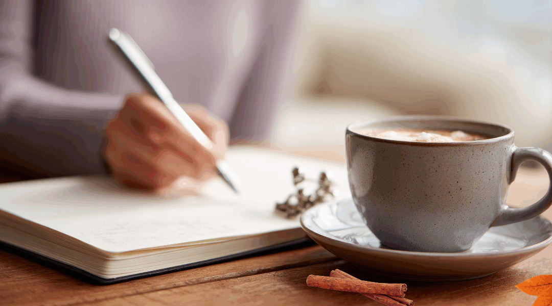 Person journaling with a warm cup of coffee beside them on a wooden table, symbolizing mindful reflection and gratitude.