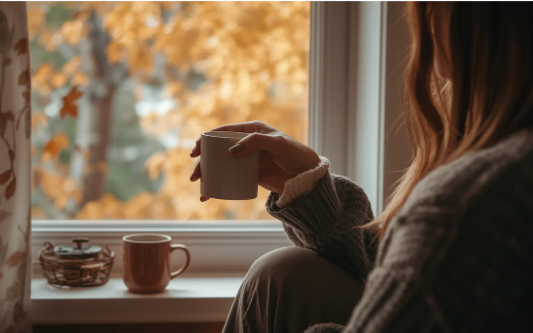 Person holding a warm drink by a window overlooking autumn trees.